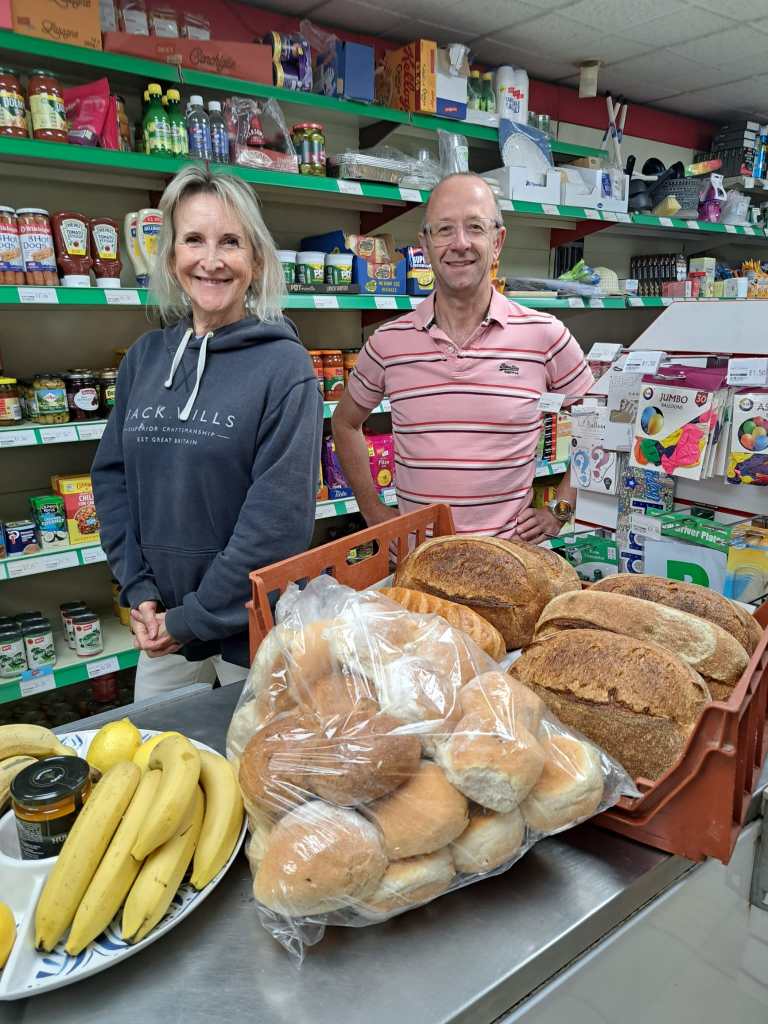 Photograph of two volunteers in the shop, standing behind some fresh bread