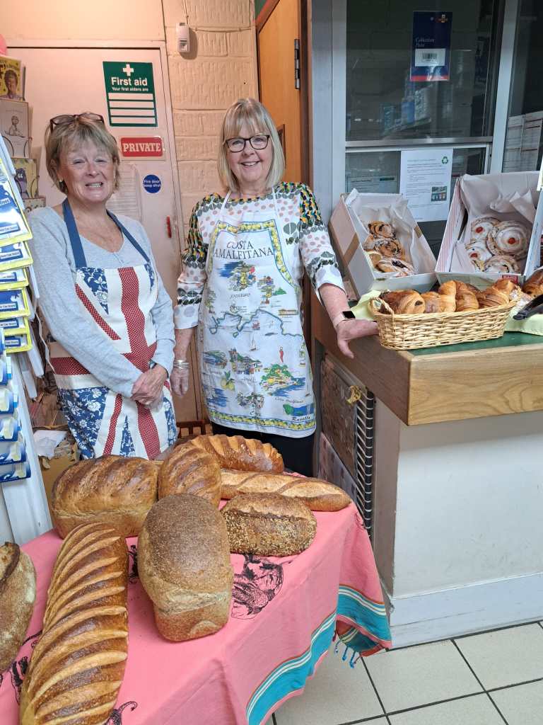 Photograph of two volunteers in the shop, standing behind some fresh bread